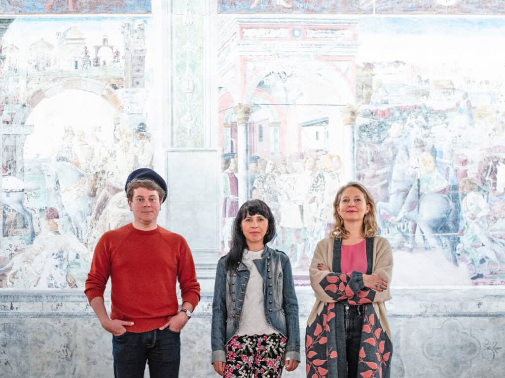 Artistic team of the exhibition “Re-Enchanting the world” in front of the murals in Palazzo Schifanoia in Ferrara, 2021; from left: Wojciech Szymański, Małgorzata Mirga-Tas, Joanna Warsza; Photo: Daniel Rumiancew